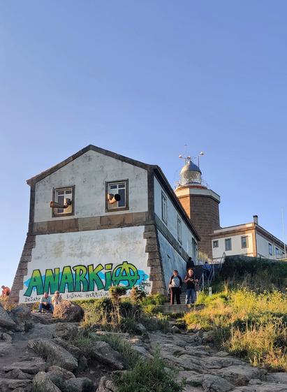 Faro de Fisterra. Delante hay un edificio en el que alguien ha dibujado  un grafiti con la palabra "Anarkia". Pueden verse varias personas alrededor del faro.