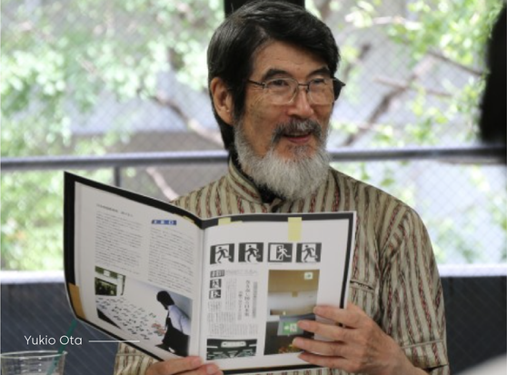 Picture of Yukio Ota 10 years ago during a presentation. He has papers in his hands depicting his work of creating the Exit Sign.