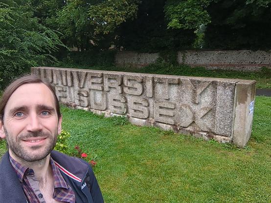 A selfie of me standing in front of a brutalist concrete monolith reading "University of Sussex" surrounded by grass and trees.