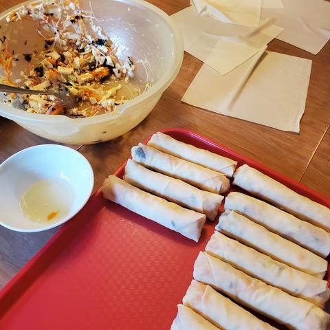 Mixing bowl with spring roll stuffing mix on far upper left. Sauce bowl with egg white in front of mixing bowl. Orange plastic cafeteria tray with uncooked spring rolls.
