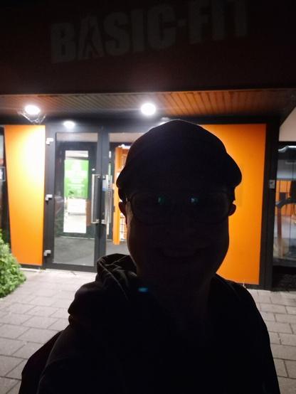 A dark silhouette of me, taking a selfie, at the entrance of the Basic-Fit in Oss. The background has more light, so I guess that's why my face appears darker