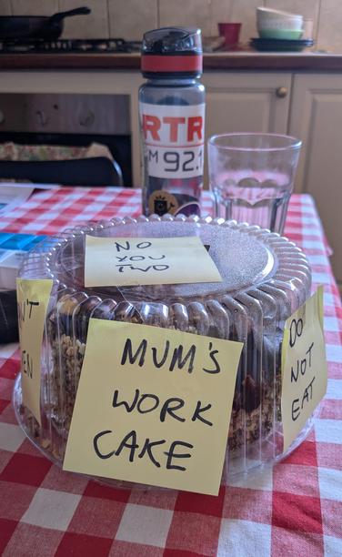A cake on a dining table with several sticky notes warning others not to eat it. Messages include MUM'S WORK CAKE, NO YOU TWO and DO NOT EAT