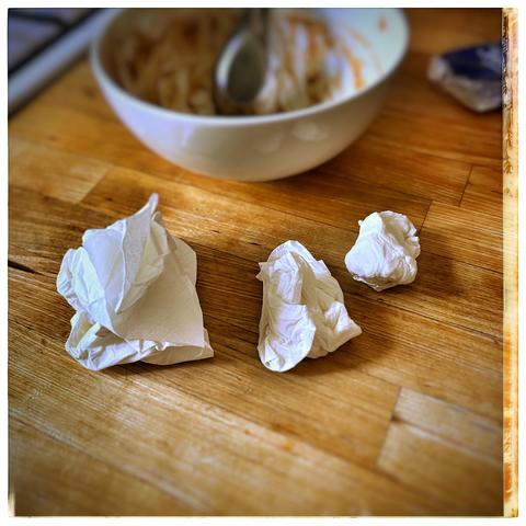 Three crumpled white paper towels on a wooden surface, with a shallow bowl of what appears to be a finished meal in the background. A metal spoon rests inside the bowl.