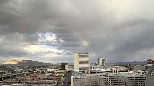 A cityscape featuring a view of buildings under a cloudy sky, with a faint rainbow visible in the background. Mountains can be seen in the distance, and a freeway runs through the landscape. First rainbow I’ve ever seen in many, many trips to Vegas.