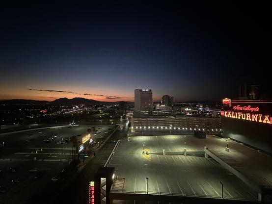 A sunrise cityscape showing the illuminated skyline with mountains in the background. Prominent features include a parking lot and a neon sign for a casino, with a gradient sunset sky transitioning from deep blue to warm orange hues.