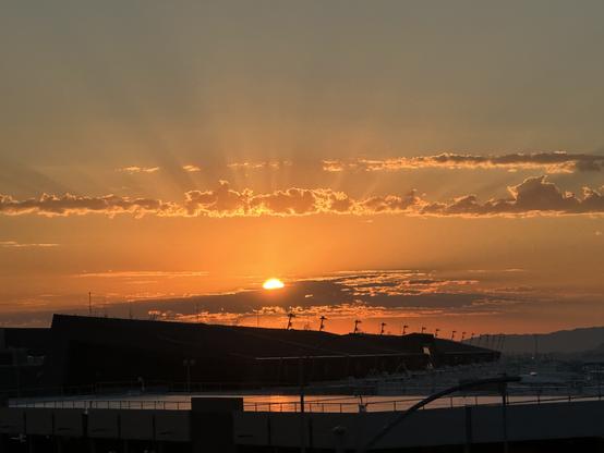 A beautiful with rays of sunlight breaking through clouds, casting a warm orange glow in the sky. In the foreground, silhouettes of a building and boats are visible against the calm water. Headed to Vegas airport.