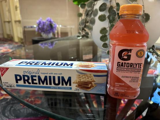 I wasn’t hung over but you wouldn’t know it from my breakfast. A box of Premium saltine crackers topped with sea salt is positioned next to a bottle of Gatorlyte, a watermelon-flavored electrolyte drink. The background features blurred floral arrangements and a patterned carpet.
