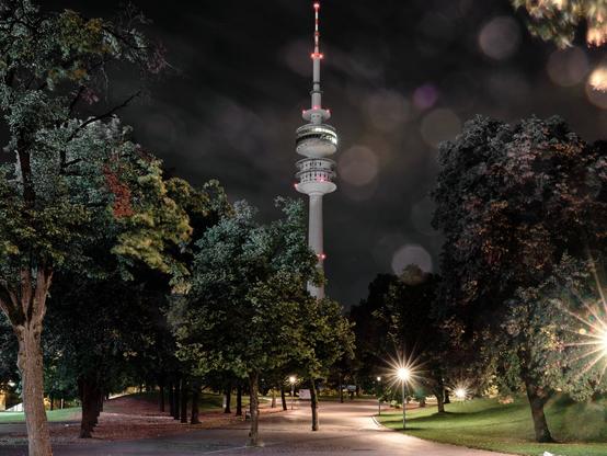 The photo you provided depicts a nighttime scene in Munich's Olympiapark. The image prominently features the Olympic Tower (Olympiaturm), an iconic landmark in Munich. The tower is illuminated, with red and white lights glowing along its structure, enhancing its visibility against the dark night sky. Surrounding the tower are various trees, adding depth and framing the image nicely. The pathway leading up to the tower is softly lit by streetlights, creating a calm and serene ambiance. Additionally, there are noticeable bokeh effects from the lights, adding a touch of artistic flair to the photograph. Overall, the image captures a peaceful nighttime atmosphere with a focus on the architectural beauty of the Olympic Tower amidst a natural setting.