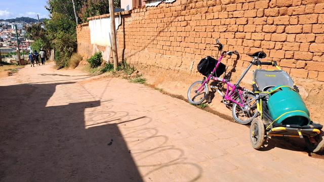 A folding bike with a trailer carrying a bottle of propane gas, leaning on a mud=brick wall in Madagascar.