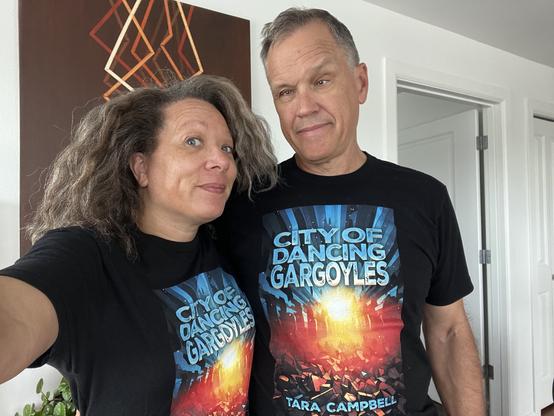 Man and woman wearing black T-shirts with blue, orange, and yellow book cover of CITY OF DANCING GARGOYLES