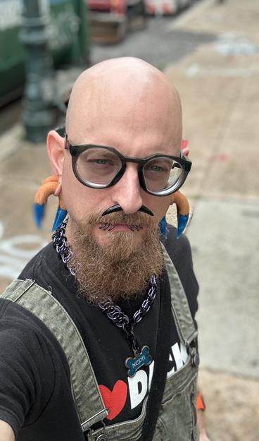 A person with a bald head and a thick full goatee poses for a selfie. They wear large glasses, a purple and black ornate chainmail collar with a blue bone shaped tag that says “NEPH”, a black t-shirt with "I ❤️ DICK" printed on it, and green overalls. Their facial accessories include a blue tiget eye stone septum tusk, 6 lip rings, and large wooden and blue resin ear hangers.