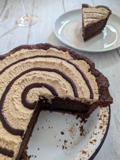 A rectangular photo of a pie, slice of the same pie and the stem of a glass on a white wooden background. The pie has a dark chocolate shortcrust. It is topped with a light-brown cream topping and a dark chocolate sauce swirl on top. The pie has been cut open to expose a mid-brown cake filling, with a layer of dark chocolate sauce at the bottom.