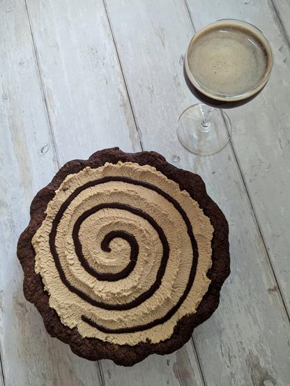 A rectangular photo taken from above of a pie and cocktail on a white wooden background. The pie has a dark chocolate shortcrust. Only the pie's topping is visible, which is a light-brown cream with a dark chocolate sauce swirl on top. The cocktail is a dark brown liquid with light brown foam on top, in a Nick and Nora glass.