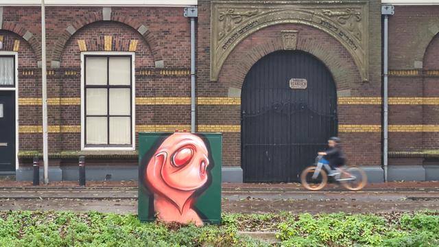 Graffiti art of a smiling earthworm on a green  utility box on the side of the road in some low vegetation in front a red brick building with two horizontal yellow brick lines and yellow accents above the window and front door. A small kid is cycling past on the sidewalk