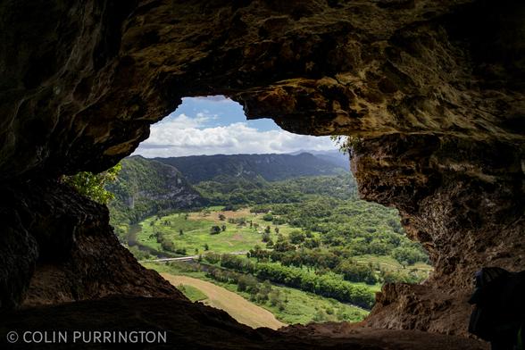 Photograph of blue sky, a mountain ridge, and a green valley with grassy, farmed areas as seen from inside a cave.