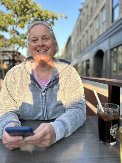 Picture of Sarah on a patio in Galt, wearing a light grey hoodie and pink t-shirt, holding her cell phone, smiling for the camera. A glass of cola is on the table beside her left elbow. A green tree is visible in the background behind her.