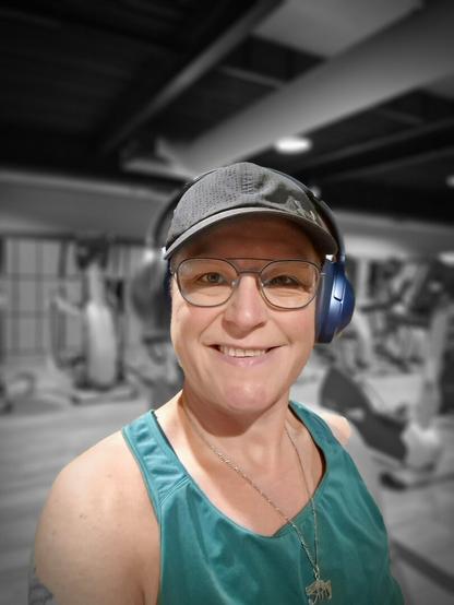 Selfie of a person riding the seated bike at the gym. Smiling at the camera. Used a filter to have me in color and the background is black and white. Wearing a green top, black cap, blue headphones and blue-rimmed glasses. In the background are several cardio machines.