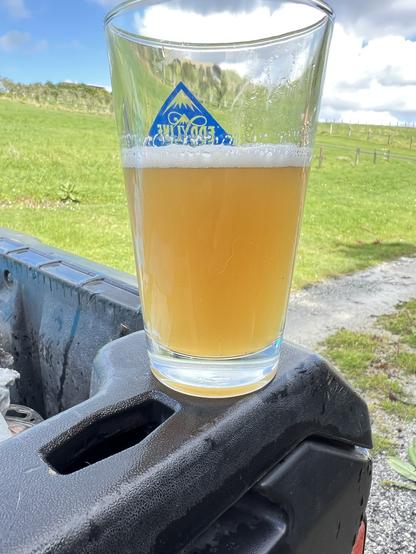 Hazy IPA in a bit more than half filled shaker pint on the rear gate of a vehicle. A bit of blue sky, clouds and green paddock in the background.