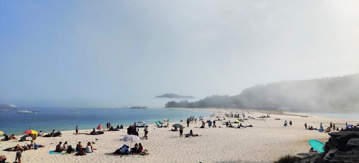 Gente en la playa, con sombrillas y en bañador. Al fondo se ve un acantilado cubierto por la niebla.
