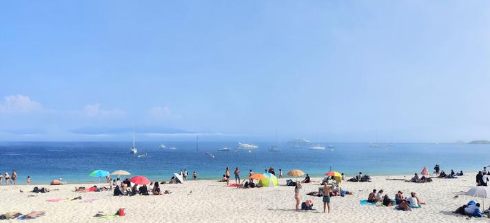 Gente en la playa con sombrillas de colores y en bañador. En el mar se ven algunos barcos y al fondo, se vislumbra la niebla.