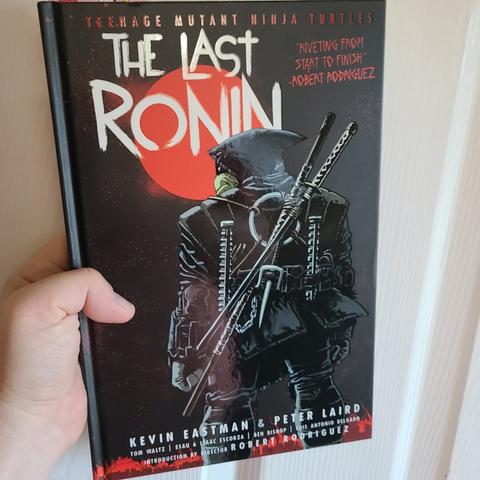 Photograph of a white person's hand holding a dark, hardcover comic book entitled "Teenage Mutant Ninja Turtles The Last Ronin." The series title is across the top and in white letters, "The Last Ronin" is shown on the left in / around a red circle. Offset to the lower right is an anthropomorphic turtle standing backwards in heavy gear with multiple weapons on their back with a black hood over their head.