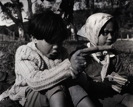 This image depicts two children in a natural outdoor setting. On the left, there is a child wearing a knitted sweater and sitting on the ground. The child appears to be holding an object made from clothes pegs, which resembles a gun due to its shape and color, with the clothes pegs arranged to simulate the barrel and handle of a weapon. On the right side of the image, there is another child with a hat on their head, also sitting on the ground, looking at the first child. The second child is holding what looks like a similar object made from clothes pegs. There are trees in the background, suggesting they might be outdoors, possibly during daylight hours due to the visibility of light and shadows.

Please note that this description is based on visible content and does not include any speculative or imaginative elements.