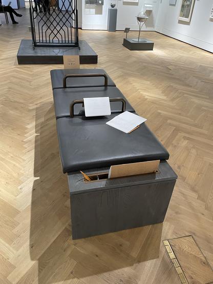 A modern bench with a dark wooden base and padded leather top is situated in a gallery space. Papers are placed on the bench, and there are openings in the sides with contents visible. The flooring is a light, herringbone pattern, and artworks