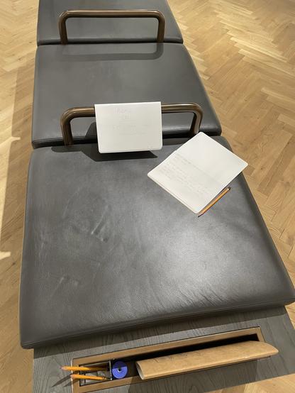 A gray padded bench with metal handles features a sign that reads "PLEASE DO SIT ON THIS INSTALLATION." Next to it, there is an open notebook with handwritten notes and a pencil. Below the bench, there is a storage area containing drawing tools