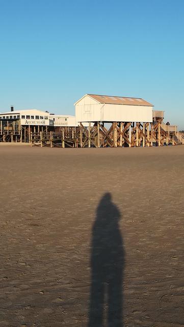 AmStrand an der Nordsee in Sankt Peter Ording. Mein länger Schatten ist im Sand zu sehen, davor eine Gruppe der berühmten Pfahlbauten.