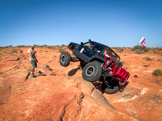 Heavily modified black cherokee XJ (with the back chopped off) lifting the front driver tire about 18". The driver's face is visible from a window with his spotter standing on the left side of the photo. Surrounded by red rock with sparse green bushes and a blue sky in the background.