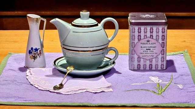 A simple & sweet tea tableau in lavender & mint green. The placemat is light purple with grass-green edging, and an embroidered Iris on the right side. In the center is a mint green tea-for-one teapot set with gilt trim and a small golden spoon, upon a pale purple handkerchief. To its left is a small, slender, vintage Swiss pitcher decorated with spiky handpainted flowers. To the right of the teapot is a purple & lilac striped box tin of Black Currant tea from Harney and Sons.