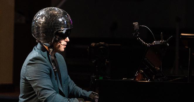 A young man wearing a blue suit, black high-tech MiMU sensor gloves and a mirrorball-like motorcycle helmet with the visor down is playing a black piano while a GoPro camera watches him from above the keyboard. This is a photograph from a performance of Laura Bowler's SHOW(ti)ME (photo credit Robin Clewley).