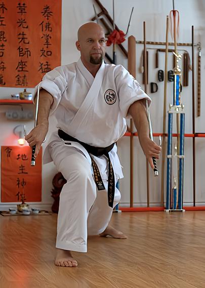 Greg in his black belt karate uniform, in barefeet, inside his dojo doing a double swords kata demonstration. Behind him are incense altars, a huge trophy & some long handled weapons for training.