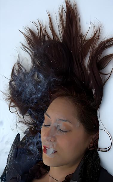 A long haired brunette woman, wearing red lipstick, black gloves & laying on the snow covered ground with her eyes closed & exhaling smoke.