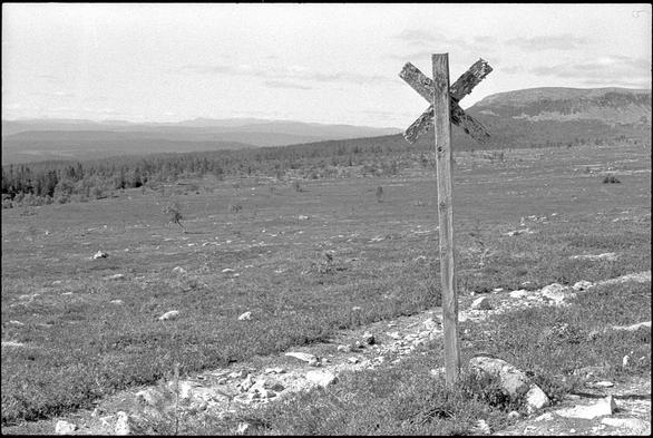 A nordic mountain landscape. In the foreground is a trail-marking in the form of a wooden cross. This particular marking is very old and weather-worn. In the distance, the lower landscape can be seen spread out. The weather is fair.