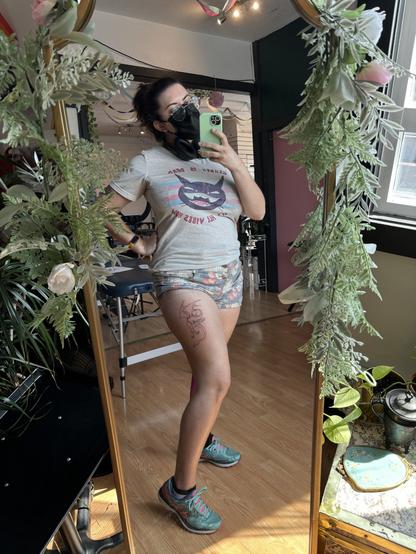 A photo of me taking a mirror selfie while on a break after completing the lines. I’m wearing a face mask, and a shirt that says “gender is dead, it’s all vibes now” with a gleeful gremlin looking character over top of a trans flag.