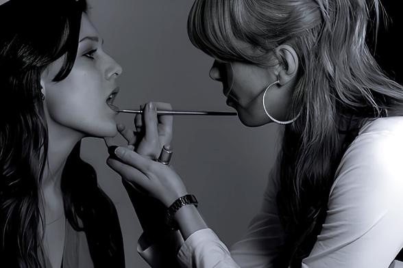 A blonde makeup artist (on right), applying lipstick on model(on left).
Black and white photo.