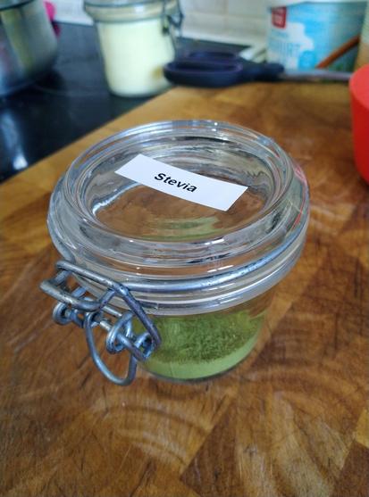 Auf einem Holzbrett steht ein Weckglas mit Metallklammer, darin befindet sich ein sehr feines, grünes Pulver, auf dem Label auf dem Deckel steht "Stevia"