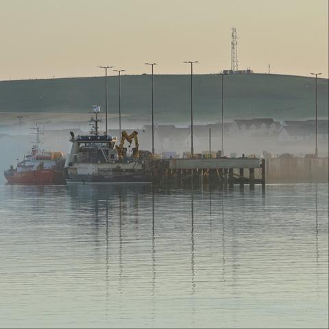 Two large boats tied up to a pier. Behind them, mist clings around the roofs of low houses, and in the distance a cellphone toward stands at the top of a low hill with warm sunrise skies behind