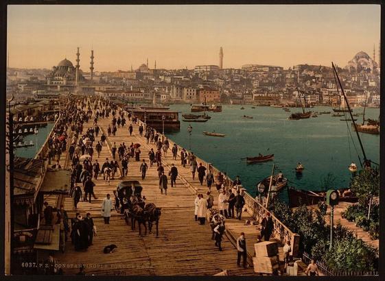 The image is a vintage postcard featuring the Galata Bridge in Istanbul, Turkey, during the period around 1890 to 1900. It shows a bird's-eye view of pedestrians walking across the bridge, with several individuals holding umbrellas. The backdrop includes minarets and mosques, providing a glimpse into the urban landscape and architectural styles prevalent during that era. The image conveys a sense of daily life in a historical city setting.