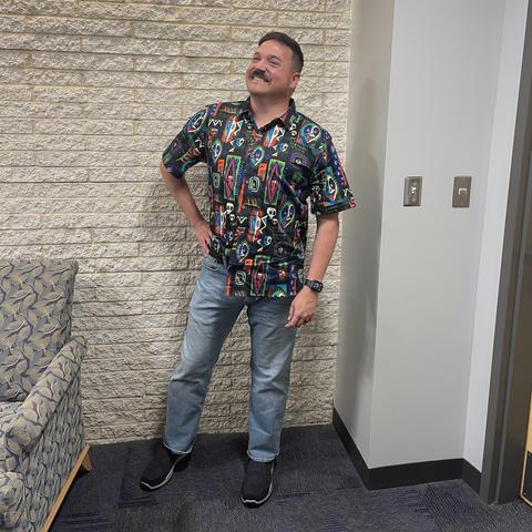 A man stands confidently with one hand on his hip, wearing a colorful patterned shirt featuring skulls and geometric designs. He is paired with light blue jeans and black sneakers. The background includes a textured stone wall and a couch.