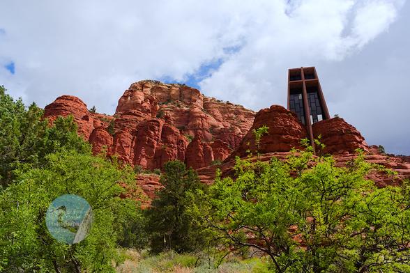 An outdoor, daylight photograph looking up, over some green shrubs to a red-hued butte or mountain with a building built into one. It emerges from a gap between to peaks and is a tall building with sides that sweep up and a facade of glass windows and a stone cross.