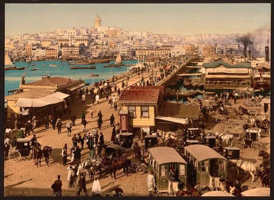 The image depicts a bustling city street in Istanbul, Turkey, likely between the years 1890 and 1900. The Galata Bridge, a significant landmark, stretches across the river, connecting different districts of the city. On either side of the bridge are rows of buildings that line the streets, suggesting a dense urban environment.

People are seen walking on the streets, engaged in various activities, which adds to the lively atmosphere of the scene. The colors are vibrant with earthy tones dominating the palette, providing a sense of the period and location.

In the background, there is a clear view of the Galata Tower, an iconic structure visible from many parts of the city. This image captures the essence of Istanbul during this era, showcasing the urban landscape and daily life of its inhabitants.