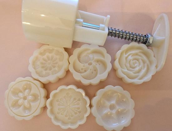 Cremefarbene Mooncake Gebäckpresse und 6 Aufsätze in verschiedenen Blütenformen - alles liegend auf einem hellrosa Untergrund.
