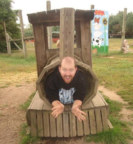 Photograph of a white, bearded, balding man inside of a wooden train steam engine toy coming out of the front of the engine car with a huge smirk on his face.