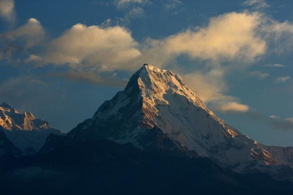 in einer nahaufnahme ist der berg annapurna south zu sehen. der gifpel wird in den frühen morgenstunden von rechts von der sonne angeleuchtet, während der rest noch im schatten liegt. im blauen himmel ist leichte, freundliche bewölkung zu sehen.