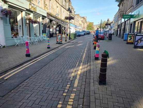 A photo of a street in the middle of a town. There are bollards all along the sides of the brick paved road and every one of them has been yarn bombed. The closest to the camera is brown with thin white stripes and a green leafy top and the others visible are multicoloured with lots of pink, yellow, blue and orange variations. A light blue cafe with chairs outside on the pavement is visible on the left hand side of the street and posters for the opticians on the right.