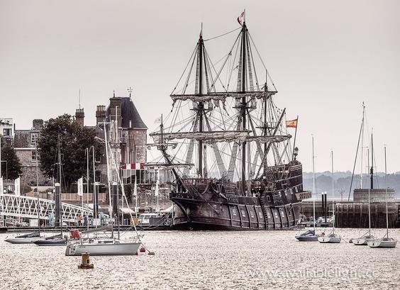 A sepia style photo of a replica of a galleon ship. The sails are down and the rigging looks very complex. It definitely has a pirate ship look about it. Moored close by is a modern small yacht and a building with a small turret is on the shore.