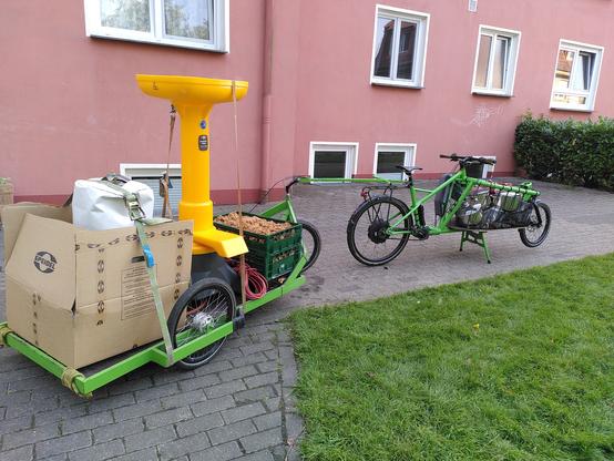 Lastenrad mit Anhänger vor einem roten Gebäude mit Fenstern. Auf dem Anhänger steht eine Kernobstmühle von Speidel, grüne Euroboxen mit Trester und ein Karton in dem die Hydropresse steht.
Im Lastenkorb des Fahrrads stehen diverse Töpfe.