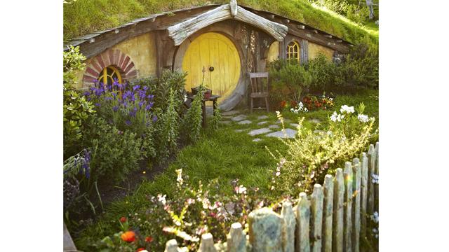Hobbiton movie set in New Zealand
A long curved-roofed home nestled into the side of a grassy green hill. It’s bright yellow door and arched front window frames make it instantly recognisable as a hobbit’s residence. 

The front garden behind a simple wooden fence is ablaze with bright colourful flowers in white, pink, red, blue, orange and yellow. A simple wooden chair rests at the front door beside a simple path made of flat smooth stones.
Image Credit: Matt Munro, Lonely Planet